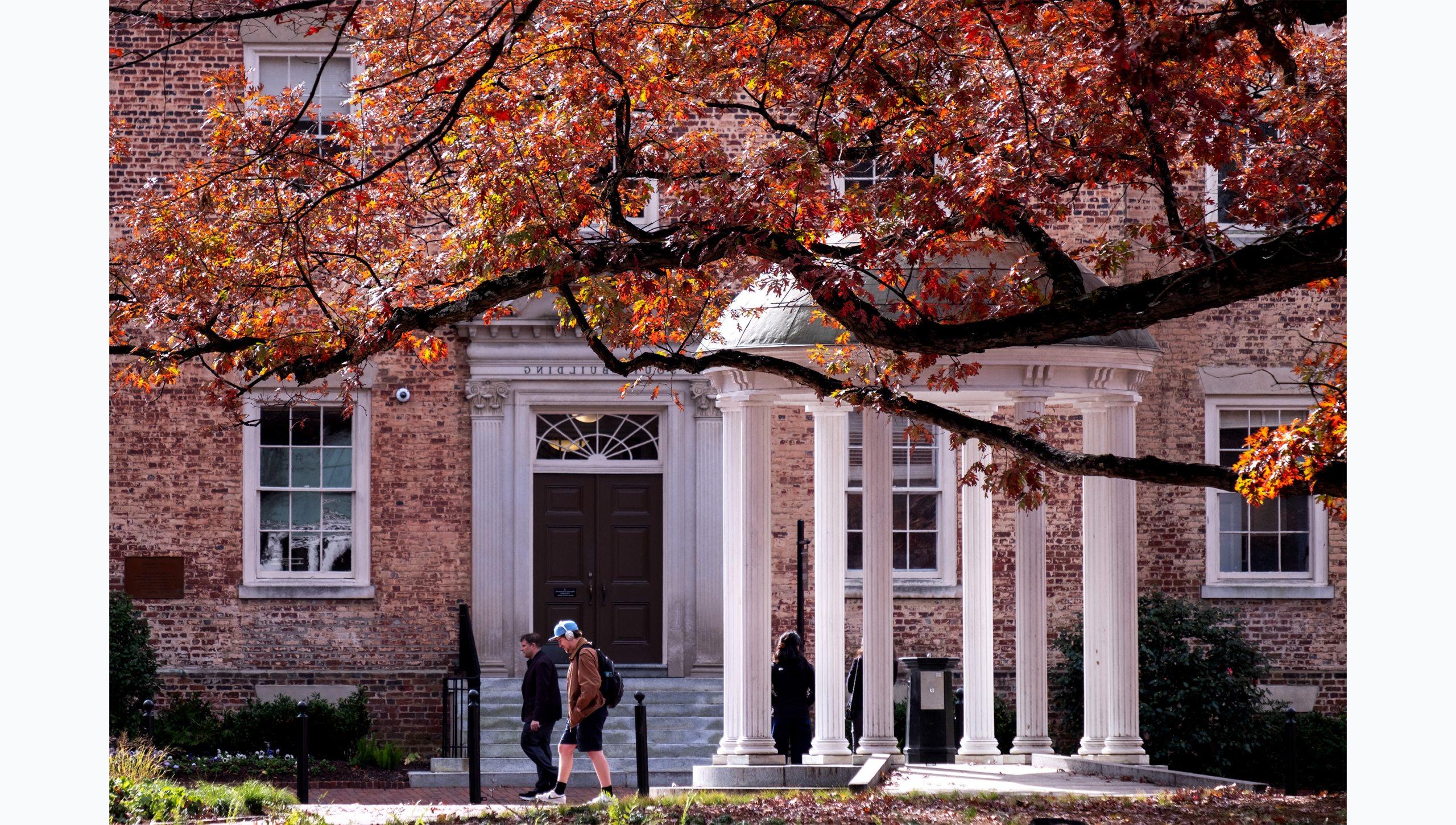 People walking by the Old Well on the campus of UNC-Chapel Hill on a fall day. Branches and yellow, red and orange leaves are seen hanging in the foreground.